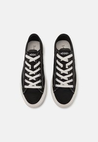 Zapatillas de lona negras con cordones blancos, costuras en contraste y suelas de goma. Presentan un logo en la lengüeta; diseño bajo clásico.
