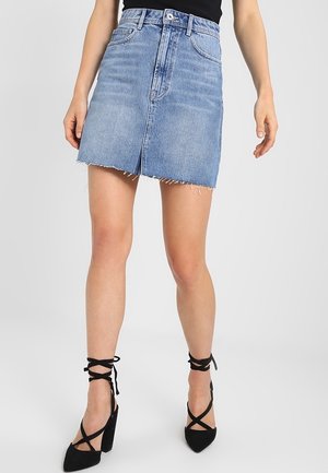 Vrouw draagt een lichtblauwe denim minirok met onafgewerkte zoom, gecombineerd met zwarte schoenen met hoge hakken en bandjes, tegen een effen achtergrond.