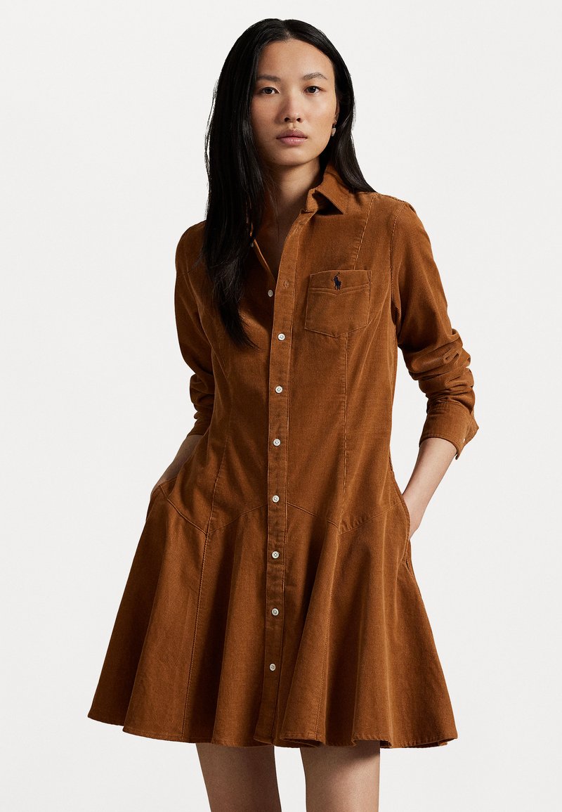 Brun kordflodress med figurnära bodice och utsvängd kjol, med knäppning fram, bröstficka och långa ärmar som är uppvikta.