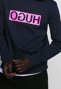 Personne portant un sweat-shirt bleu marine avec un logo rectangulaire rose affichant le mot inversé "HUGO" sur la poitrine.