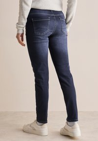 Donkerblauwe spijkerbroek met een slim fit, voorzien van twee achterzakken, contrasterend wit stiksel en een gladde denimtextuur. Gecombineerd met witte sneakers.