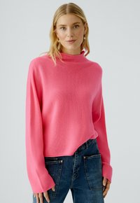 Rosa Pullover mit hohem Kragen, weichem Strickstoff und weiten Ärmeln, kombiniert mit dunkelblauen Jeans, die einen lässigen Look kreieren.