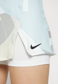 Falda atlética de color azul claro con patrones geométricos abstractos en gris y blanco. Presenta un logotipo de Nike en negro cerca del dobladillo, acompañada de pantalones cortos blancos.