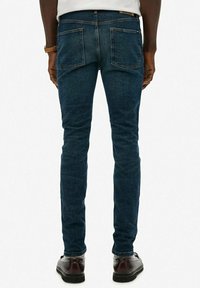 Hombre vestido con jeans ajustados de color azul oscuro, camisa blanca, zapatos de cuero marrón y un reloj beige, de pie de espaldas contra un fondo liso.