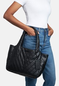 Kvinna i vitt linne och blå jeans som håller en stor svart kviltad läderhandväska med en tagg på kedjan.