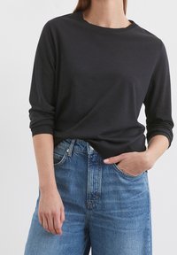 Žena oblečená v černém tričku s dlouhým rukávem, mírně zastrčeném do vysokých modrých džínsů, s jednou rukou v kapse.
