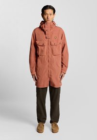 Veste légère à manches longues corail avec capuche, deux poches avant, fermetures à pression et tissu texturé. Associée à un pantalon olive et des chaussures beiges.