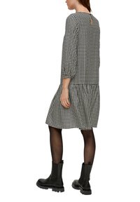 Czarna i biała sukienka w wzór houndstooth z długimi rękawami, plisowanym dołem, detalem w kształcie klucza z tyłu, noszona z czarnymi botkami za kostkę i przezroczystymi rajstopami.