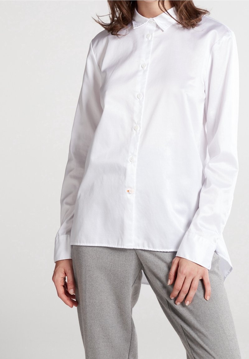 Chemise blanche à manches longues en coton lisse, avec un col, une fermeture à boutons et un ourlet légèrement arrondi. Associée à un pantalon gris.