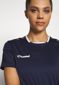 Jonge vrouw met haar naar achteren gebonden, draagt een marineblauw hummel sportshirt met witte accenten op een effen lichte achtergrond.