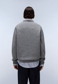 Maglione grigio a maglia con colletto alto e orlo a coste, indossato su una camicia a righe blu e bianche, mostrato da dietro.
