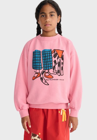 Fille aux cheveux tressés portant un sweat-shirt rose avec une illustration de chien et de jambes, ainsi qu'un pantalon rouge à motifs, sur fond blanc.