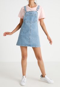 Jasnoniebieska sukienka ogrodniczka z regulowanymi ramiączkami, kieszenią z przodu i dopasowanym fasonem. Zestawiona z jasnoróżową koszulką i białymi trampkami.