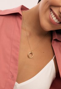 Goldene Halskette mit ineinander verschlungenen runden Anhängern und einem pinken Akzent. Getragen über einem weißen Oberteil, gegen ein pinkes Hemd.