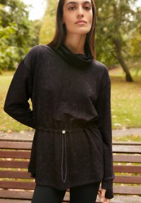 Čierny svetr s kapucňou a jemným vzorom, so šnúrkou v páse a dlhými rukávmi. Mäkkučký materiál, voľný strih a otvory na palce v manžetách.