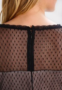 Une personne blonde porte un haut en dentelle noire transparente avec des pois et une fermeture éclair qui descend le haut du dos.