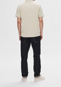 Polo beige avec col, manches courtes et design uni, associé à un jean bleu foncé et des chaussures de couleur claire.