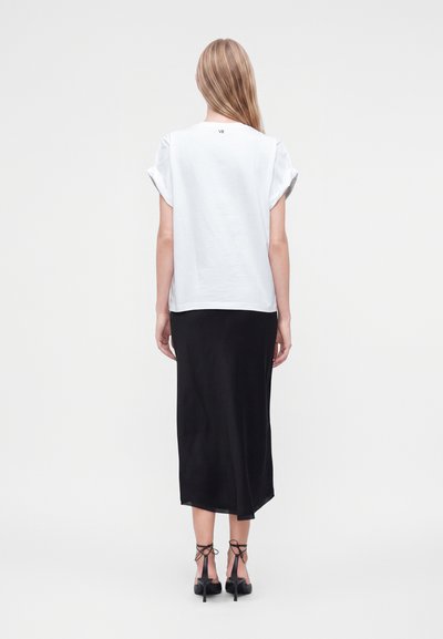 Woman with long blonde hair wearing a white short-sleeve t-shirt, black midi skirt, and black heeled sandals, shown from the back.