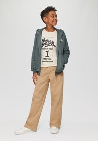 Grüne Zip-Hoodie über einem beigen Grafik-T-Shirt mit schwarzem Text, kombiniert mit hellbraunen weiten Hosen und weißen Sneakers.