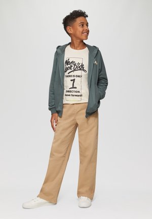Groene ritsvest over een beige graphic T-shirt met zwarte tekst, gecombineerd met tan wijde broek en witte sneakers.