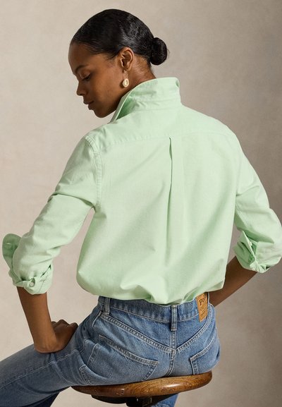 Vrouw met donker haar in een knot, draagt een lichtgroene blouse met knopen en blauwe spijkerbroek, zit op een houten krukje, met de rug naar de kijker en kijkt opzij.