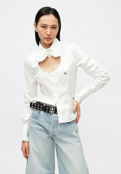 Camisa branca de manga comprida com gola franzida, decote recortado e detalhe bordado. Combinada com jeans azul claro desgastados e um cinto preto.