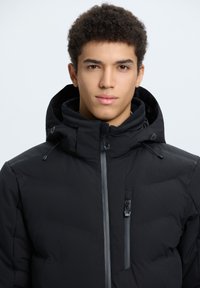 Giubbotto piumino nero con collo alto e cappuccio regolabile. Presenta una tasca a zip verticale e dettagli in zip grigia a contrasto. Tessuto liscio.