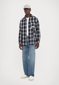 Rutet flanellskjorte i marineblå og hvit, med en frontlomme, kombinert med lyseblå jeans og hvite sneakers. Grå caps fullfører looken.