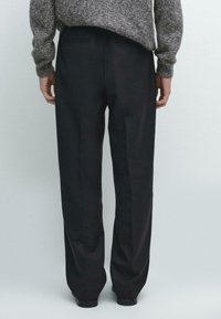 Pantalon noir ample en tissu texturé, avec une ceinture lisse et des détails de poche arrière. Vue arrière affichée.