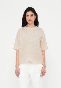 Beige strikket sweater med korte ærmer og rund halsudskæring, der har en afslappet pasform og en blød tekstur, kombineret med hvide hørbukser.
