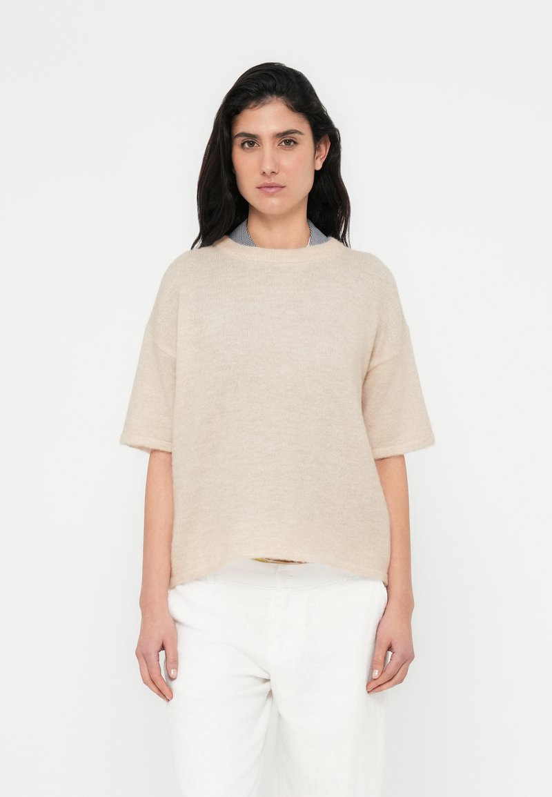 Beige strikket sweater med korte ærmer og rund halsudskæring, der har en afslappet pasform og en blød tekstur, kombineret med hvide hørbukser.