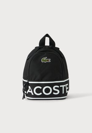 Sac à dos noir Lacoste avec bande logo blanche et emblème de crocodile vert, poche avant zippée et poignée de transport supérieure.