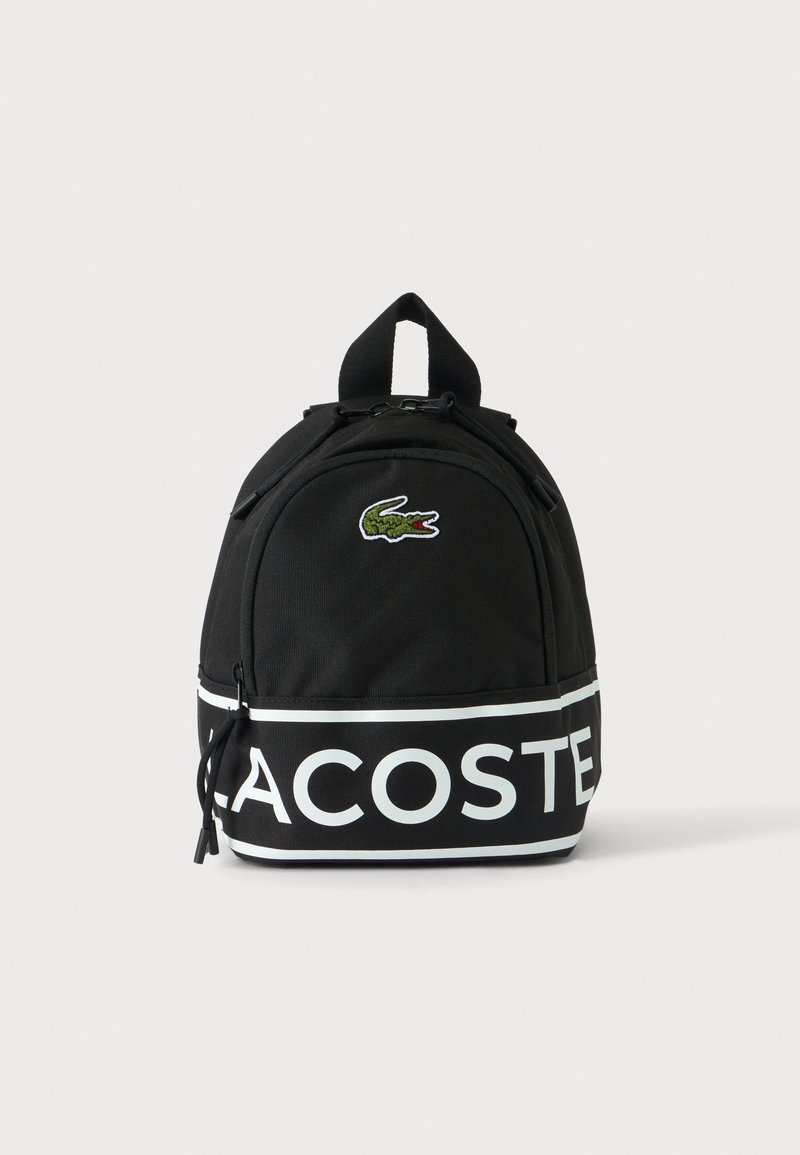 Sac à dos noir Lacoste avec bande logo blanche et emblème de crocodile vert, poche avant zippée et poignée de transport supérieure.