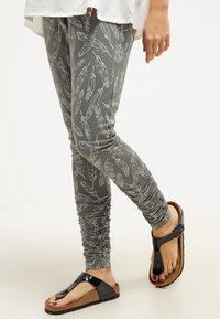 Šedé legíny s bílým vzorem peří, elastickou texturou a nařasenými manžetami, v kombinaci s černou a hnědou sandálou.