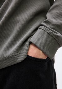 Άτομο που φοράει γκρι μπλούζα με υφή και μακριά μανίκια, με το χέρι στην τσέπη μαύρου  παντελονιού, εμφανιζόμενο σε κοντινό πλάνο του κορμού και του χεριού.