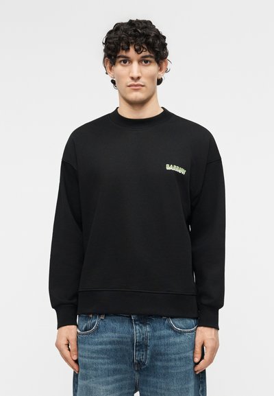 Νεαρός άνδρας με σγουρά μαλλιά που φοράει μαύρο φούτερ με το λογότυπο "Barrow" στο στήθος και μπλε τζιν, στέκεται μπροστά σε απλό φόντο.