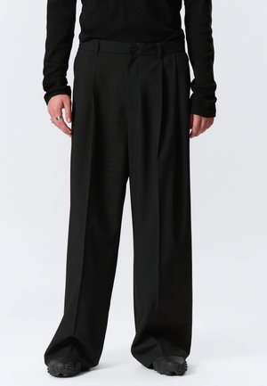 Persoană purtând pantaloni negri largi cu pliuri, bluză neagră cu mâneci lungi și pantofi negri texturati, stând în picioare pe un fundal simplu.