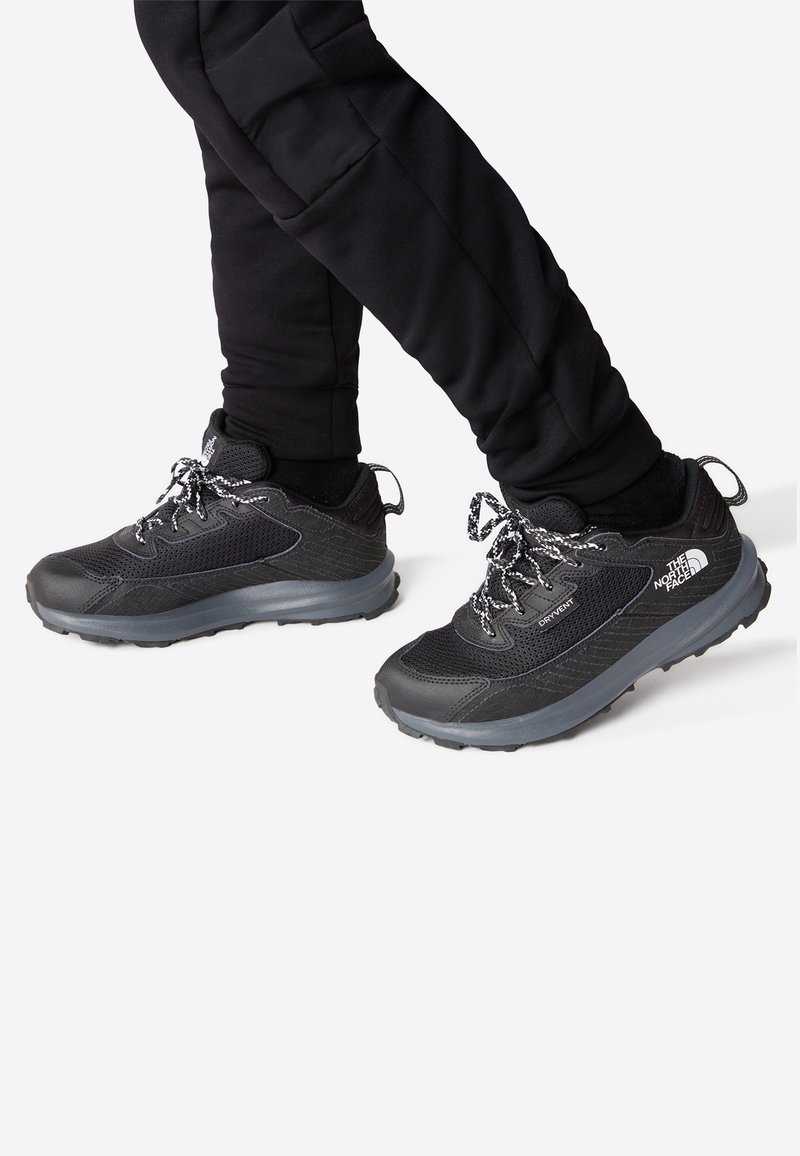 Baskets de sport noires en matière synthétique et en maille, avec une surface texturée, des lacets noirs avec des accents blancs et de épaisses semelles en caoutchouc gris.