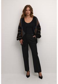 Cardigan a maglia con strisce nere, marroni e grigie, abbinato a pantaloni neri a vita alta e tacchi a punta.
