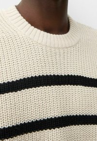 Suéter de punto en color crema con dos rayas horizontales negras. Presenta un cuello redondo acanalado y un patrón de tela texturizado.