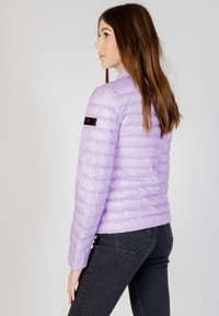 Helle lila Puffjacke mit gestepptem Design, taillierter Passform und schwarzem Branding-Detail am linken Arm. Kombiniert mit dunklen Jeans.