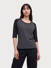 Joven mujer con una camiseta de manga raglán gris oscuro y pantalones cargo negros, de pie frente a un fondo liso y claro.
