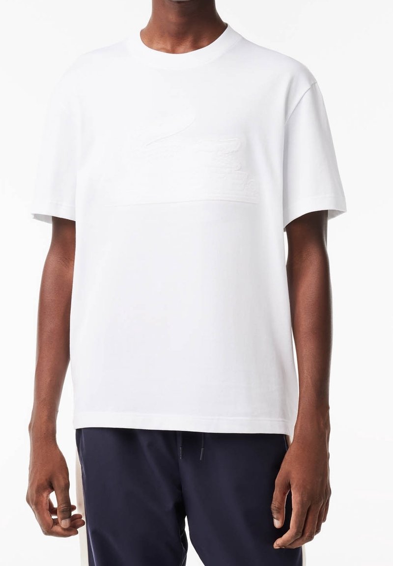 Homme portant un T-shirt à col rond blanc uni à manches courtes avec un logo discret en relief sur la poitrine et un pantalon bleu marine.