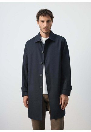 Man wearing a dark navy blue buttoned coat over a white shirt and brown pants, standing against a plain light background.