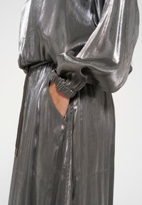 Metallisches Silber, langärmliges Kleidungsstück mit elastischen Manschetten und Bund. Verfügt über eine Fronttasche; weiche, glänzende Textur; subtile vertikale Streifen.