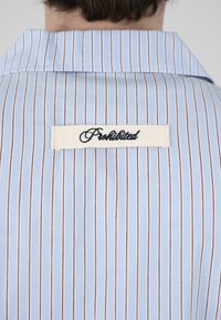 Vue de dos d'une personne portant une chemise à rayures bleu clair avec un patch blanc brodé « Interdit » sur le haut du dos.