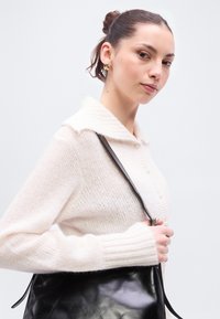 Ελεφαντόδοντο πλεκτή ζακέτα με απαλό, υφασμένο φινίρισμα, διαθέτει γιακά και λεπτομέρεια με κουμπί, φορεμένη με μαύρη δερμάτινη τσάντα ώμου.