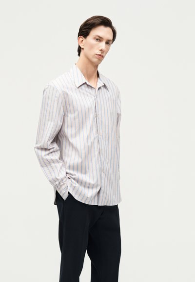 Νέος άνδρας που στέκεται με τα χέρια στις τσέπες, φορώντας μακρυμάνικο πουκάμισο με κατακόρυφες ρίγες και μαύρο παντελόνι, μπροστά σε απλό ανοιχτό φόντο.