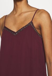 Femme portant un top sans manches violet foncé avec des fines bretelles et une bordure décorative en perles le long de l'encolure et du bord supérieur.