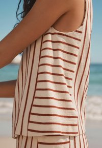 Sleeveless top in cream with bold rust stripes. Textured, relaxed fit with a curved hem. Material appears lightweight and summery.
